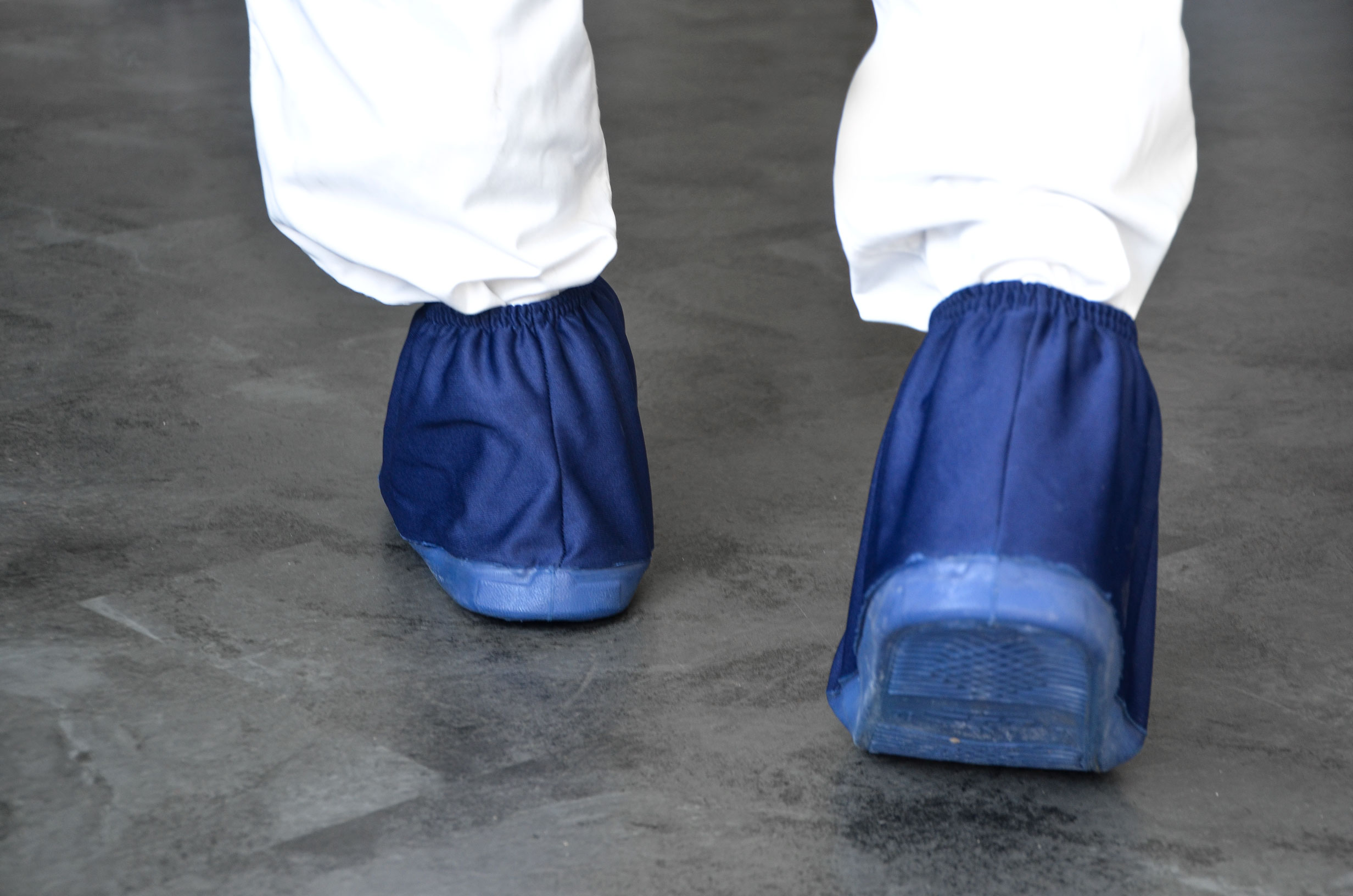 Person mit weißen Arbeitshosen trägt blaue Überschuhe als hygienischen Schuhschutz auf grauem Boden