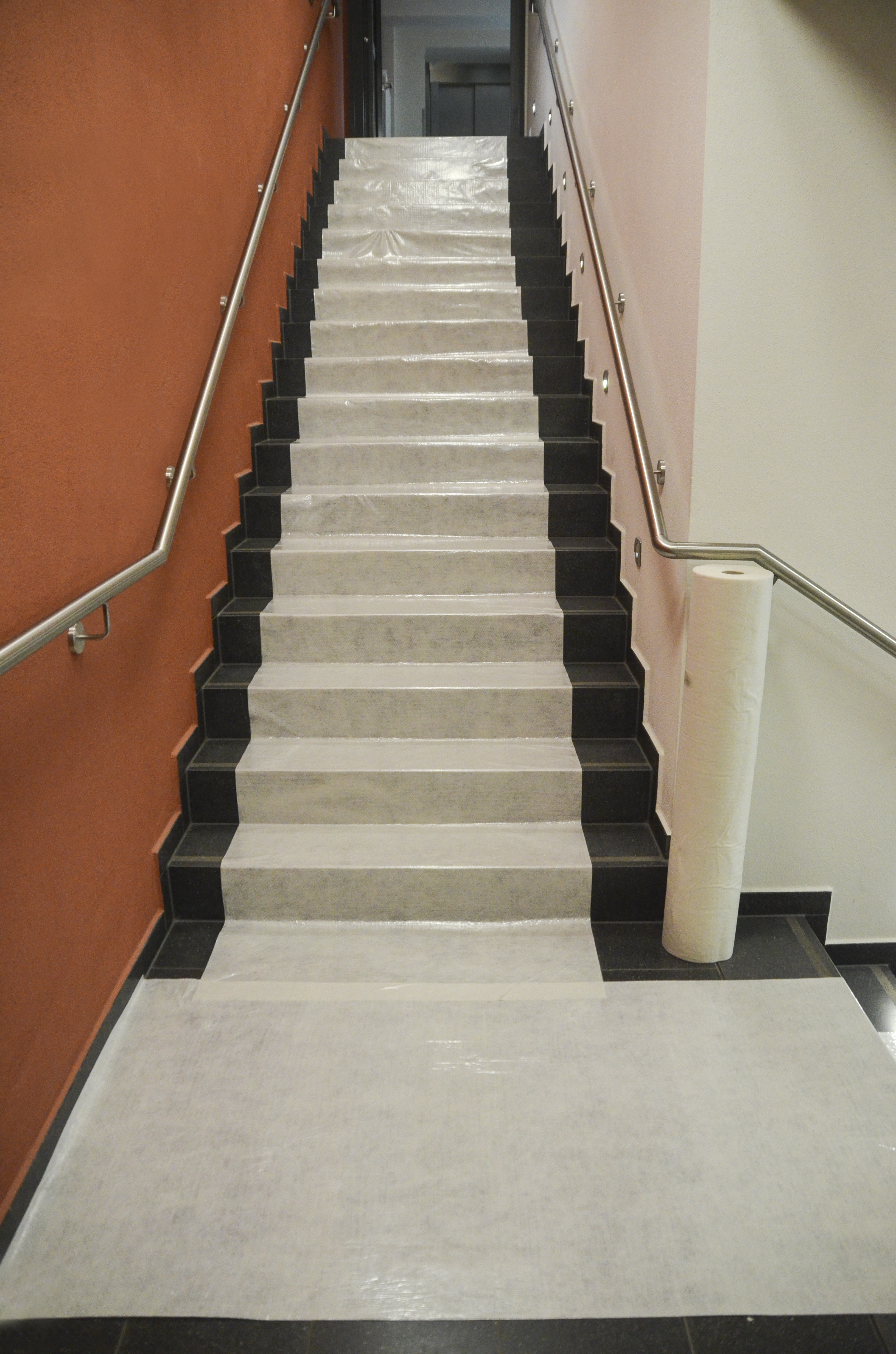 Mit Treppenschutzvlies vollständig abgedeckte Treppe mit schwarzem Rand in einem Treppenhaus, rechts daneben eine Rolle des Abdeckvlieses