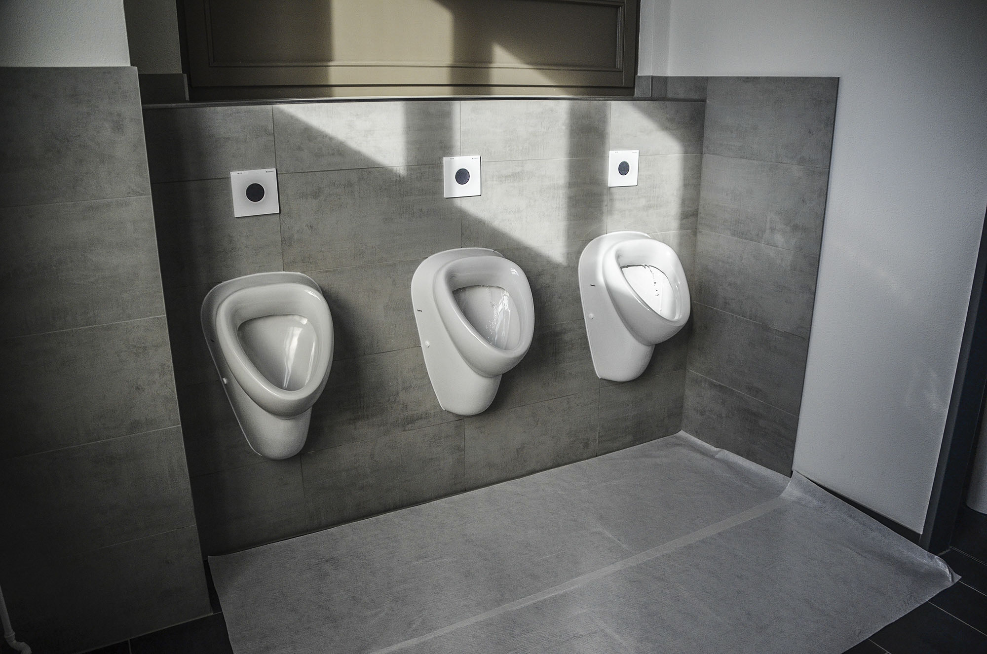 Drei nebeneinander montierte Wandurinale in einer modernen, gefliesten Herrentoilette mit hygienischer Bodenabdeckung