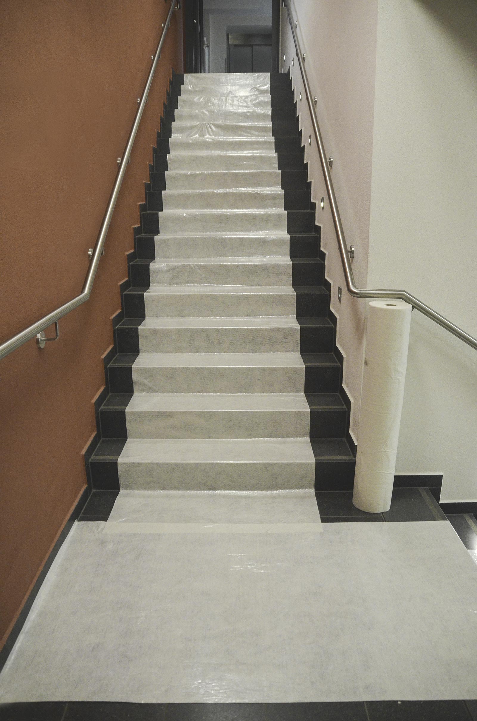 Treppe im Innenbereich mit verlegter selbstklebender Schutzfolie und daneben stehender Rolle zum Abdecken von Stufen während Bau- und Renovierungsarbeiten
