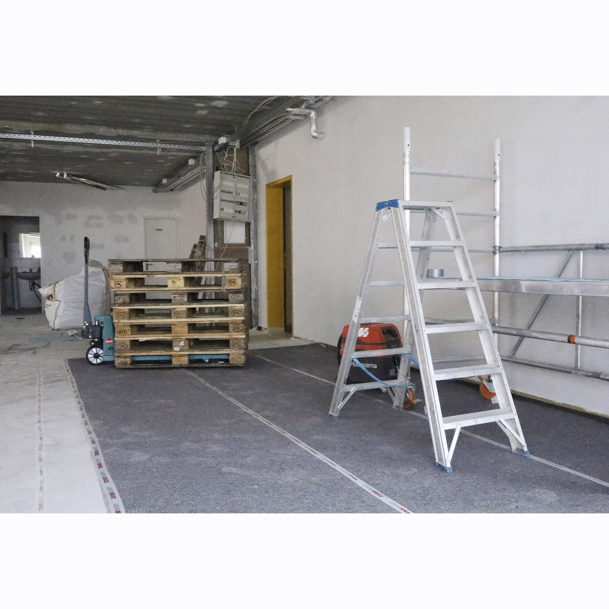 Baustellenszene in einem Innenraum mit Aluminium-Stehleiter, gestapelten Holzpaletten, Gerüst und Baugeräten auf vorbereitetem Boden