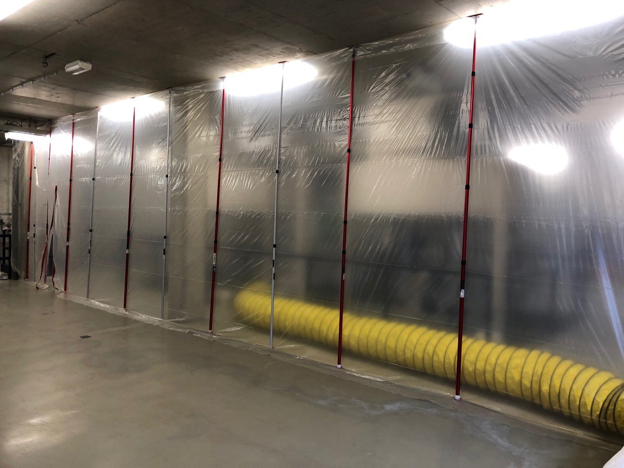 Transparente Staubschutzwand aus Folie mit roten Teleskopstangen und gelbem Lüftungsschlauch in einer Baustellenhalle