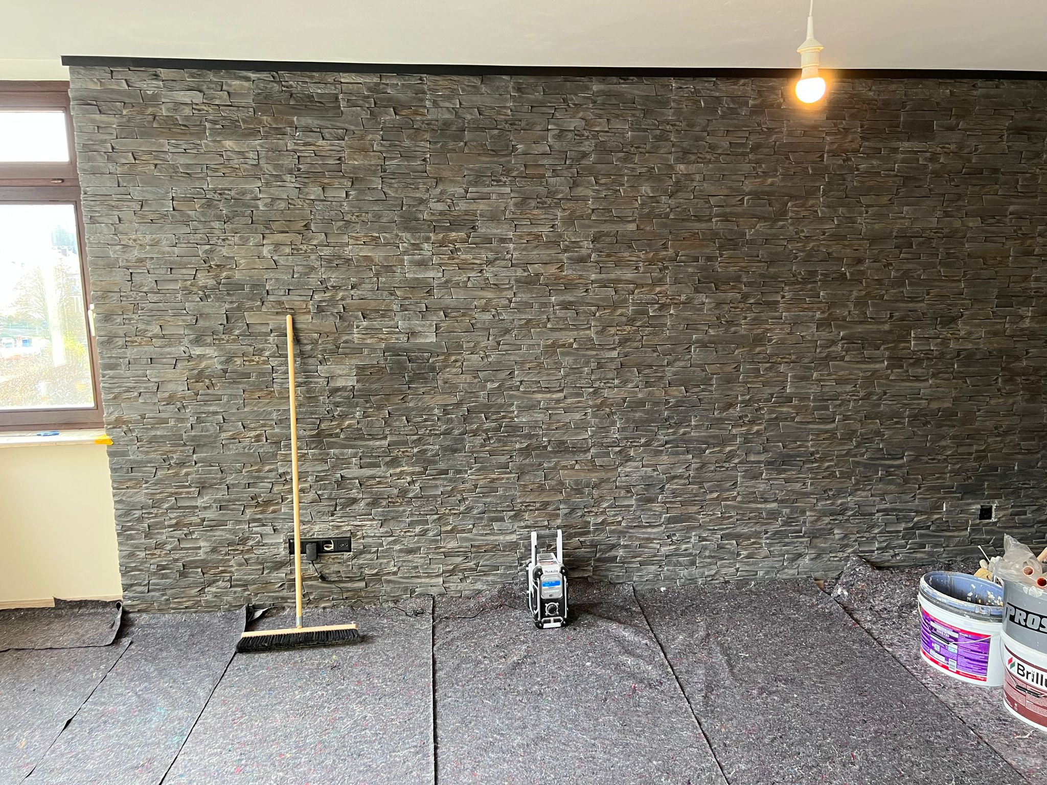 Wohnzimmer im Ausbau mit großflächiger grauer Natursteinwand, Malervlies auf dem Boden sowie Besen, Eimer und Werkzeug vor der Wand