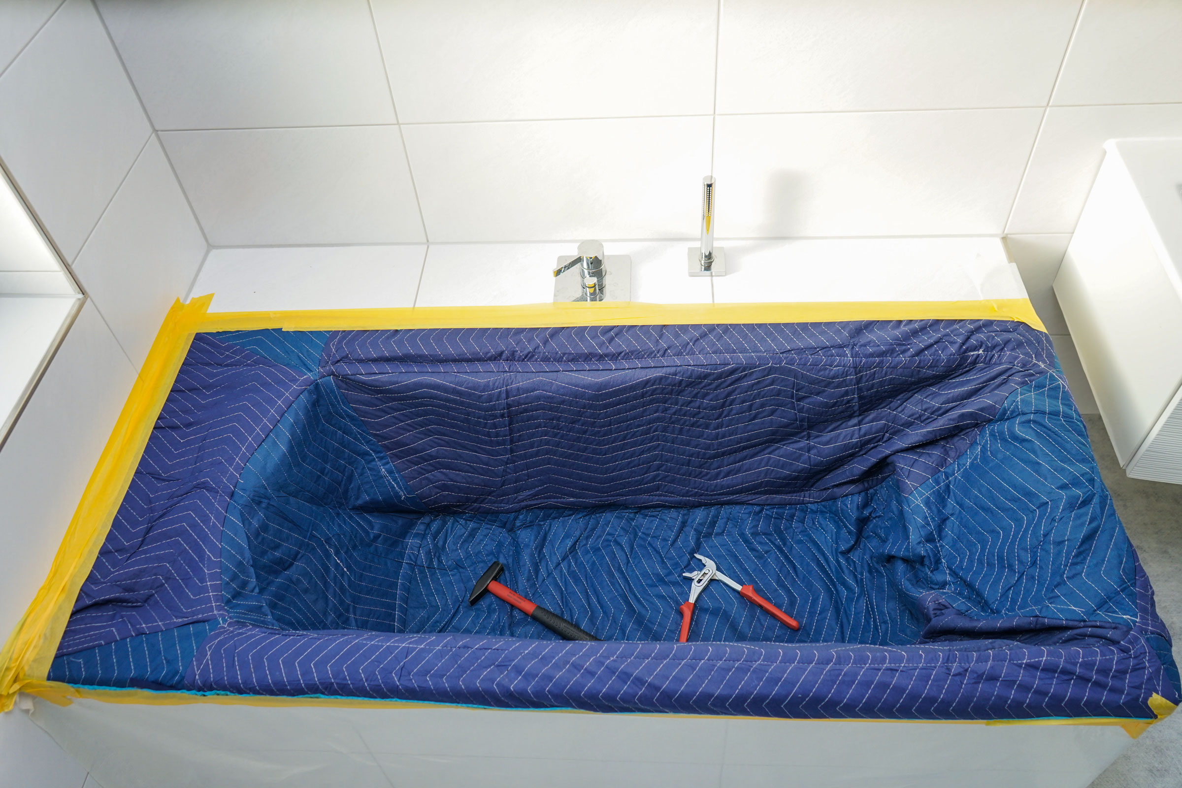 Badewanne mit blauer Schutzdecke und abgeklemmten Rändern, vorbereitet für Renovierung, mit Hammer und Zange im Wanneninneren