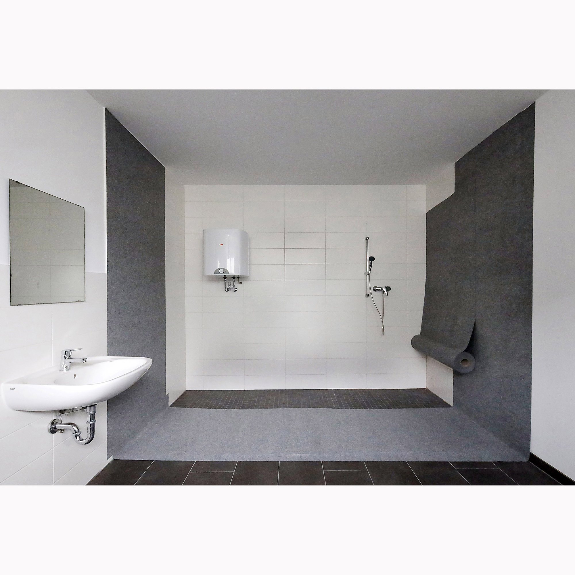 Geräumige, barrierefreie Dusche mit grauer fugenloser Boden- und Wandabdichtung, weißem Waschbecken, Wandspiegel, Durchlauferhitzer und Handbrause im modernen Bad