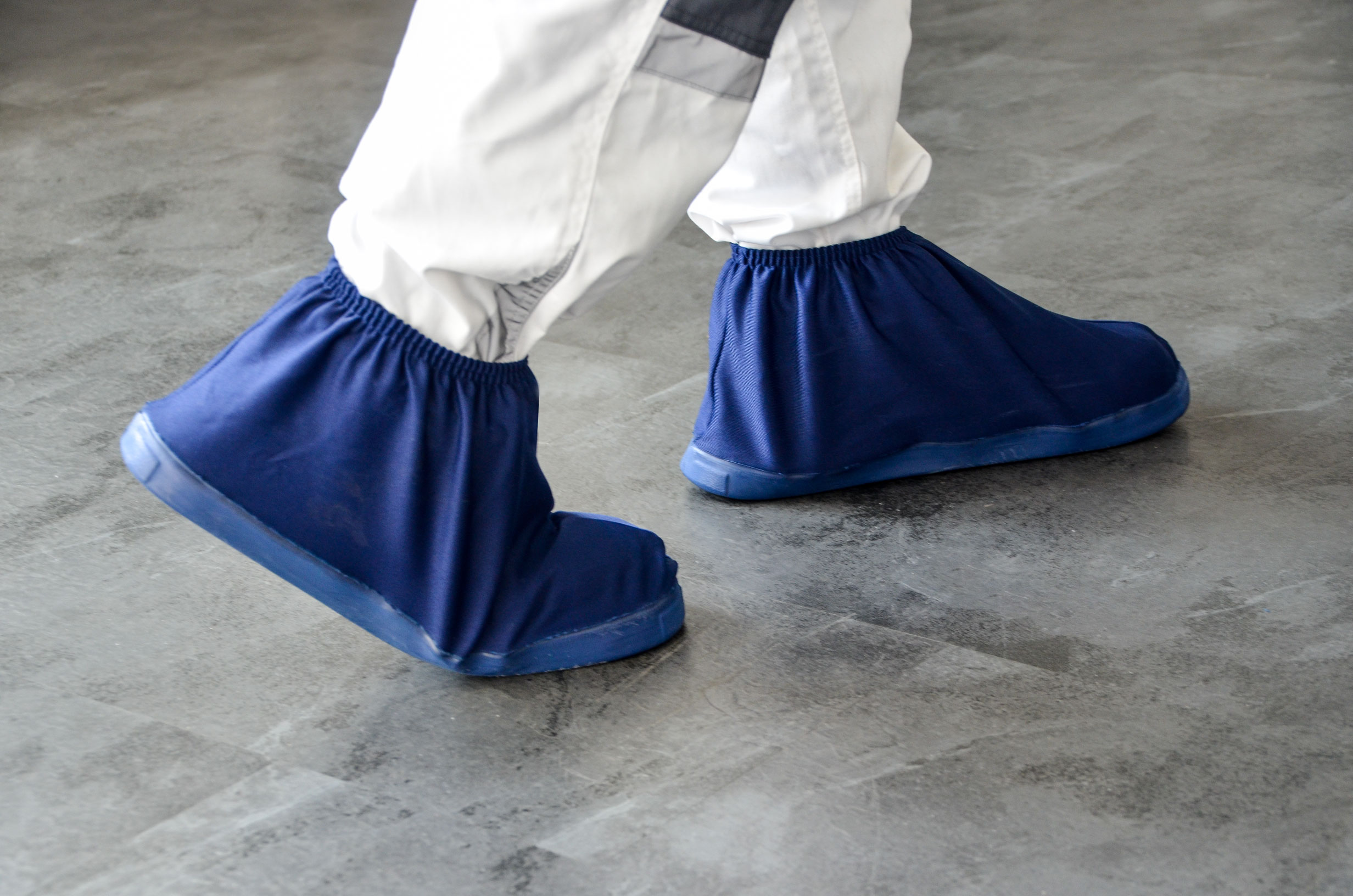 Person trägt blaue Überschuhe als rutschhemmenden Hygieneschutz über Arbeitsschuhen auf grauem Boden