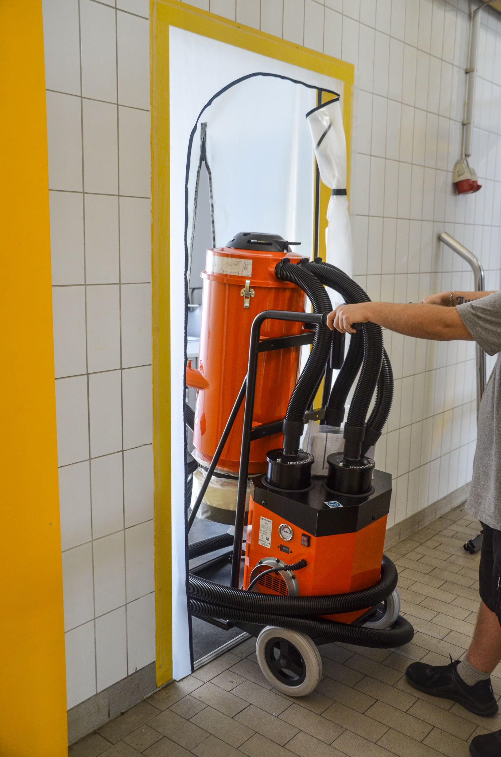 Mitarbeiter schiebt einen orangefarbenen Industriesauger mit mehreren Schläuchen durch eine provisorische Staubschutzöffnung in einem gefliesten Raum