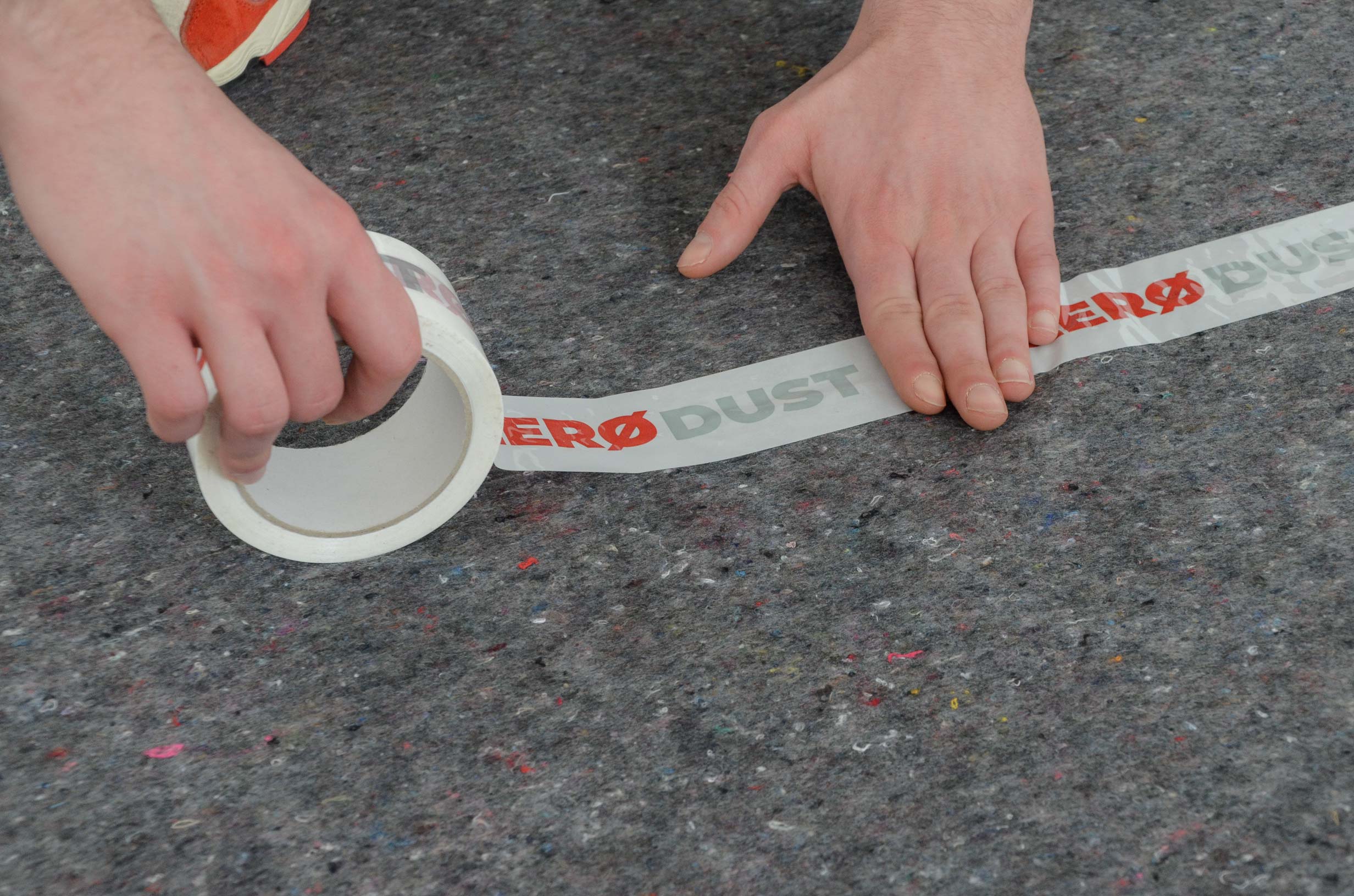 Hände rollen ZERO DUST Klebeband auf einem grauen Abdeckvlies aus, um den Boden beim Renovieren staubfrei zu schützen