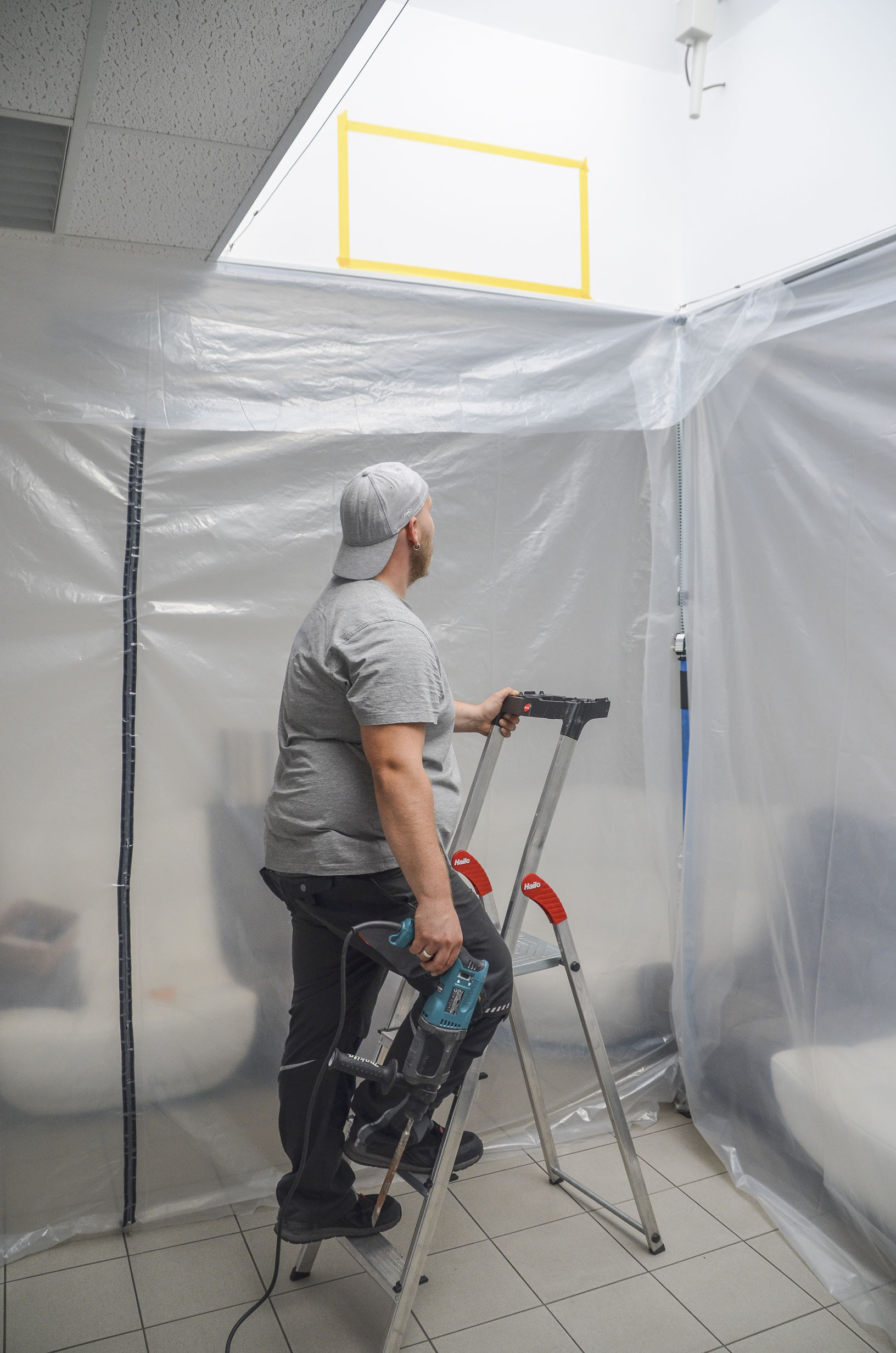Handwerker steht auf einer Leiter in einem mit Folie staubdicht abgetrennten Innenraum und hält einen elektrischen Bohrhammer für Deckenarbeiten