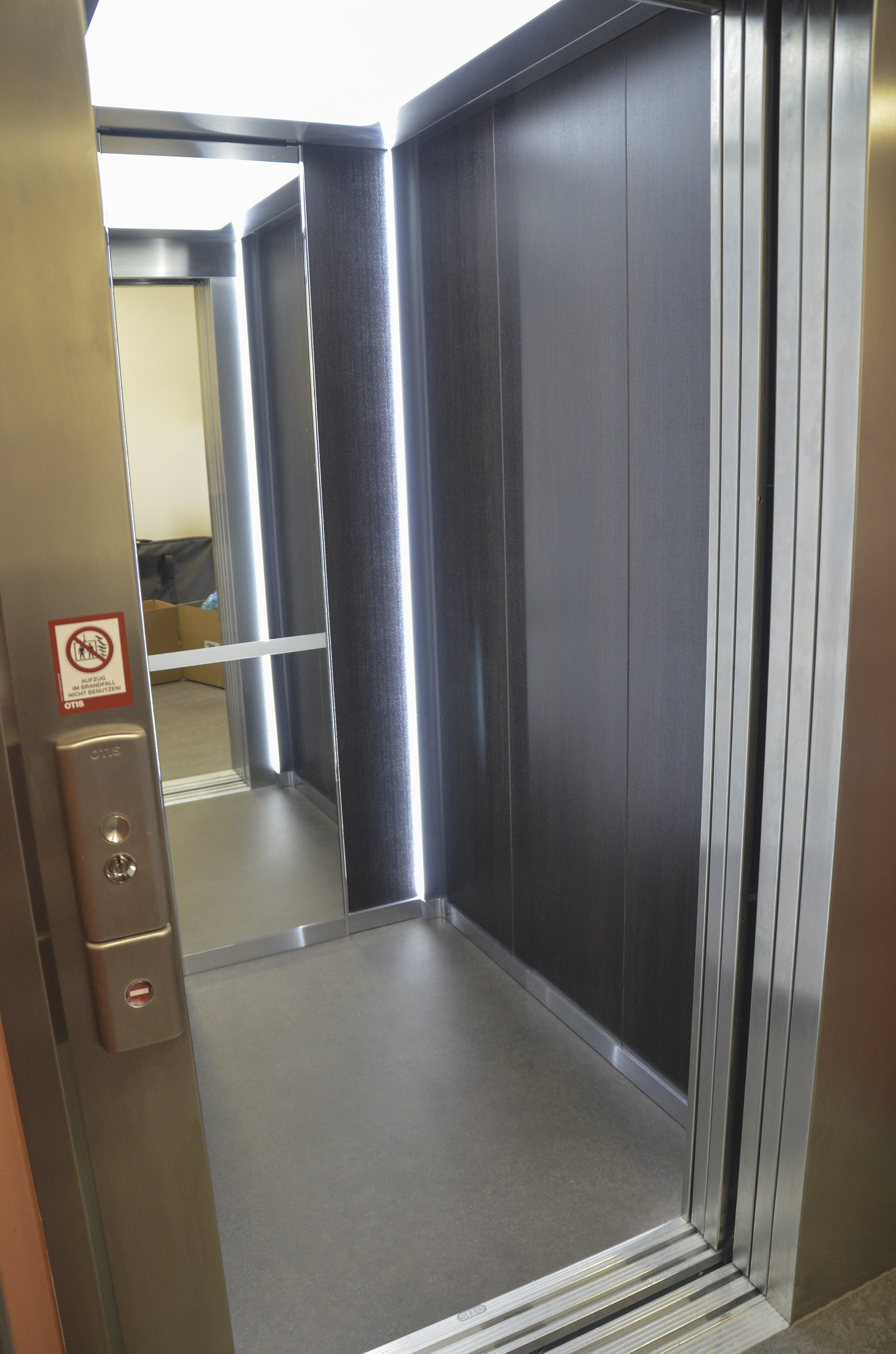 Geöffneter moderner Personenaufzug mit dunklen Wandpaneelen, LED-Lichtleisten, Edelstahlrahmen und Bedientableau