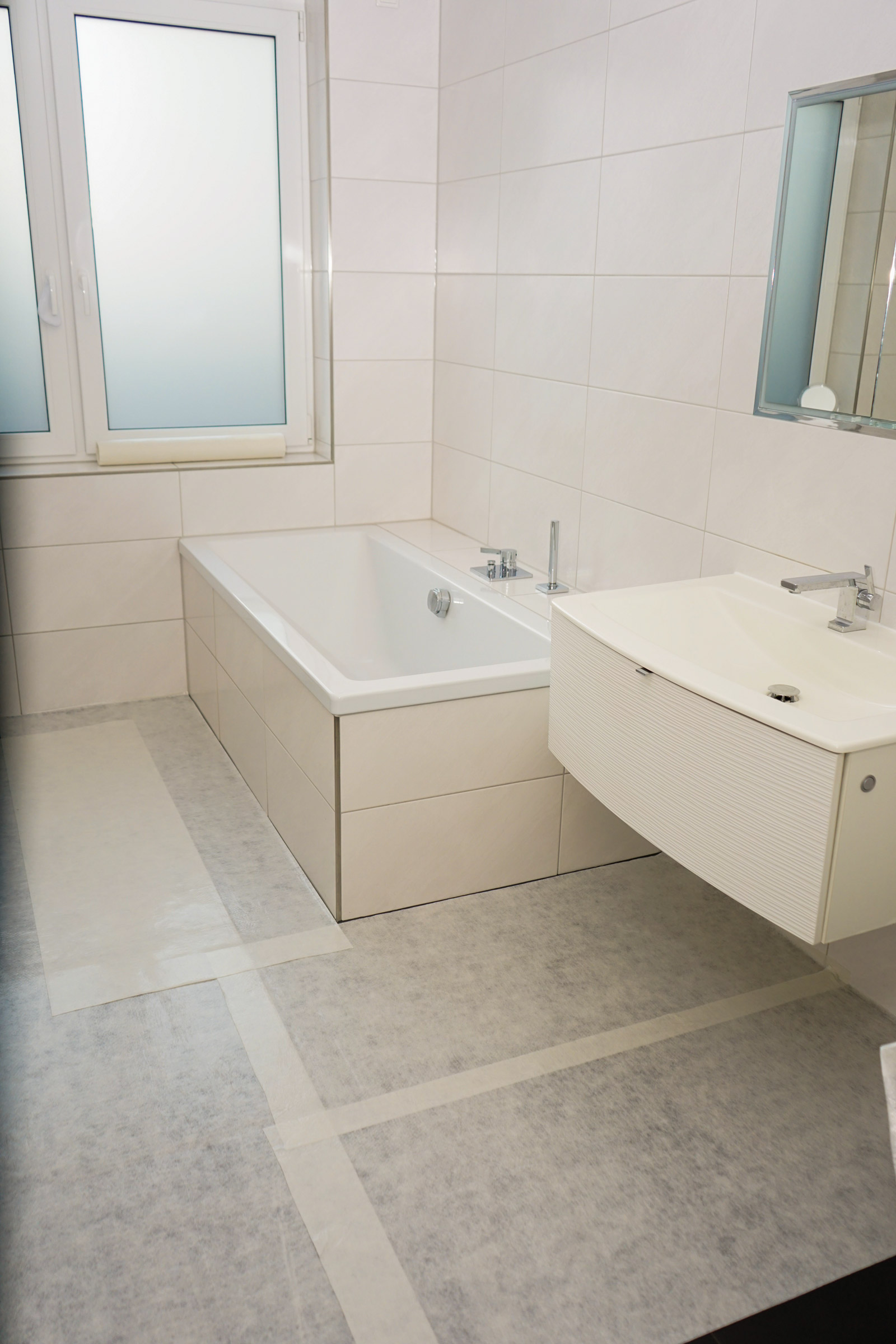 Helles Badezimmer mit rechteckiger Einbau-Badewanne, Waschtisch mit Armatur, gefliestem Wänden und mit Schutzvlies abgedecktem Boden während Renovierungsarbeiten