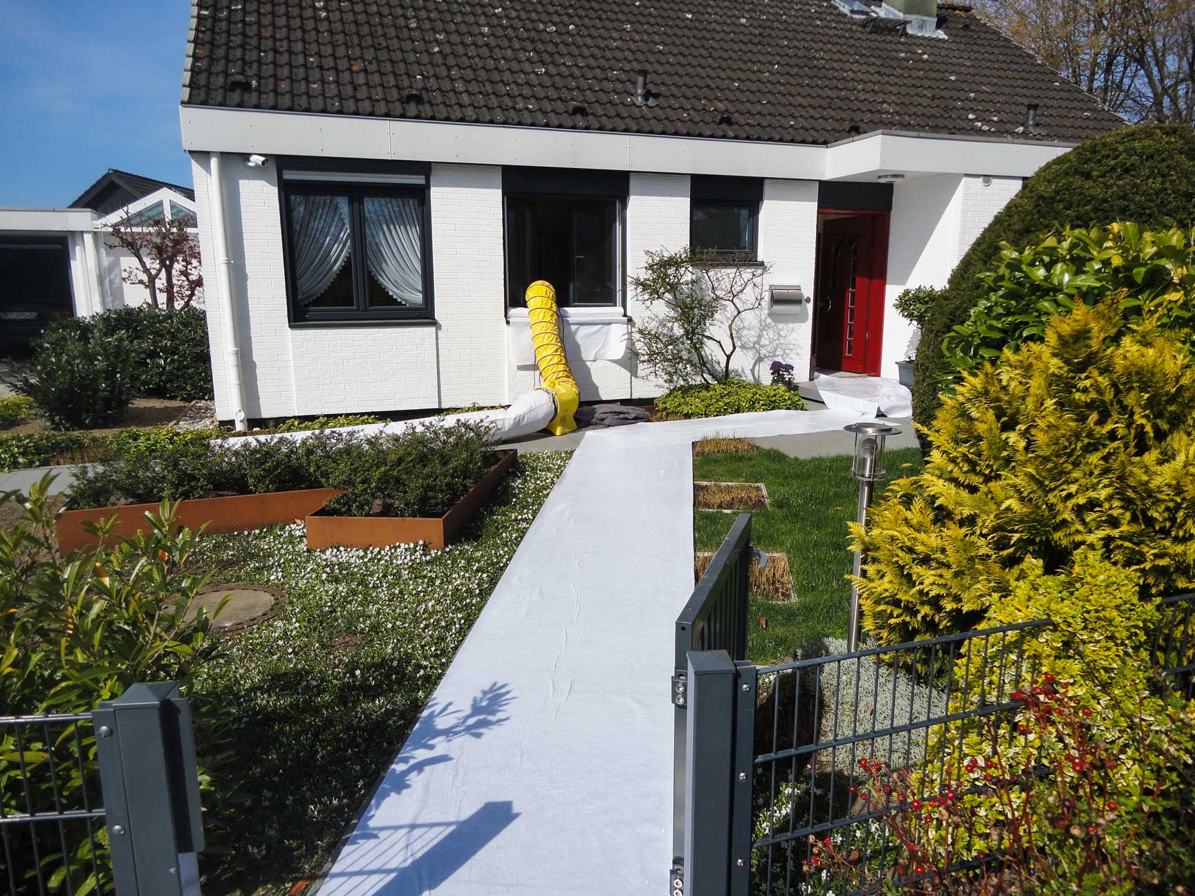 Einfamilienhaus mit weißer Fassade, abgedecktem Weg im Vorgarten und gelbem Lüftungs- oder Trocknungsschlauch am Fenster