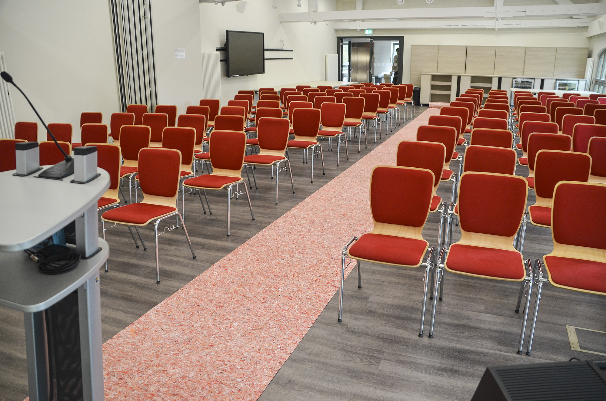 Reihenweise aufgestellte rote Konferenzstühle in einem hellen Tagungsraum mit Rednerpult und Bildschirm im Hintergrund