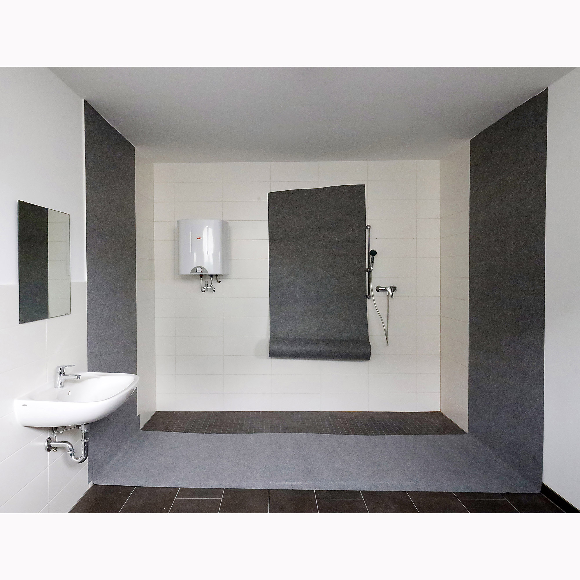 Barrierefreies Badezimmer mit bodengleicher Dusche, grauer Wand- und Bodenverkleidung, weißem Waschbecken, Spiegel, Durchlauferhitzer und Handbrause