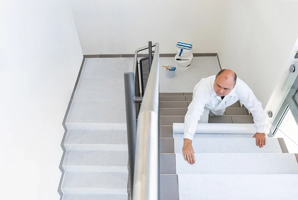 Handwerker verlegt weißes Treppenschutzvlies als Abdeckung auf Stufen in einem Treppenhaus, Malerwerkzeug steht im Hintergrund