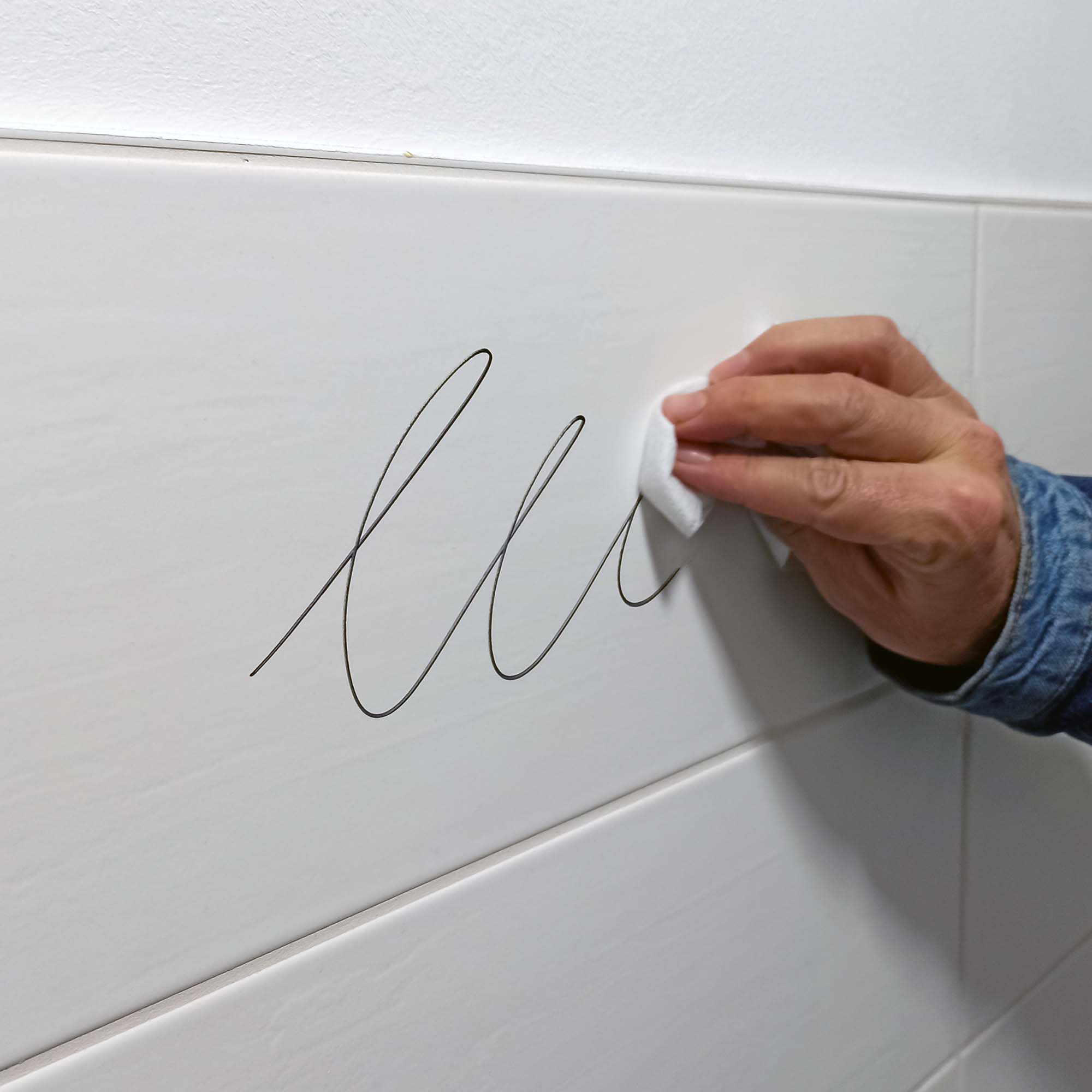 Nahaufnahme einer Hand, die mit einem Tuch schwarze Filzstiftlinien von einer weißen Fliesenwand wischt und die leicht zu reinigende Oberfläche zeigt