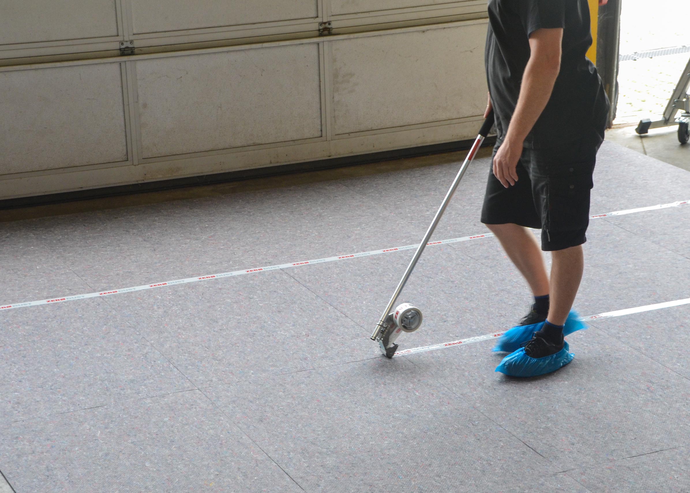 Person mit Handabroller und Teleskopstiel bringt Klebeband auf einem textilen Bodenbelag an und trägt blaue Überschuhe