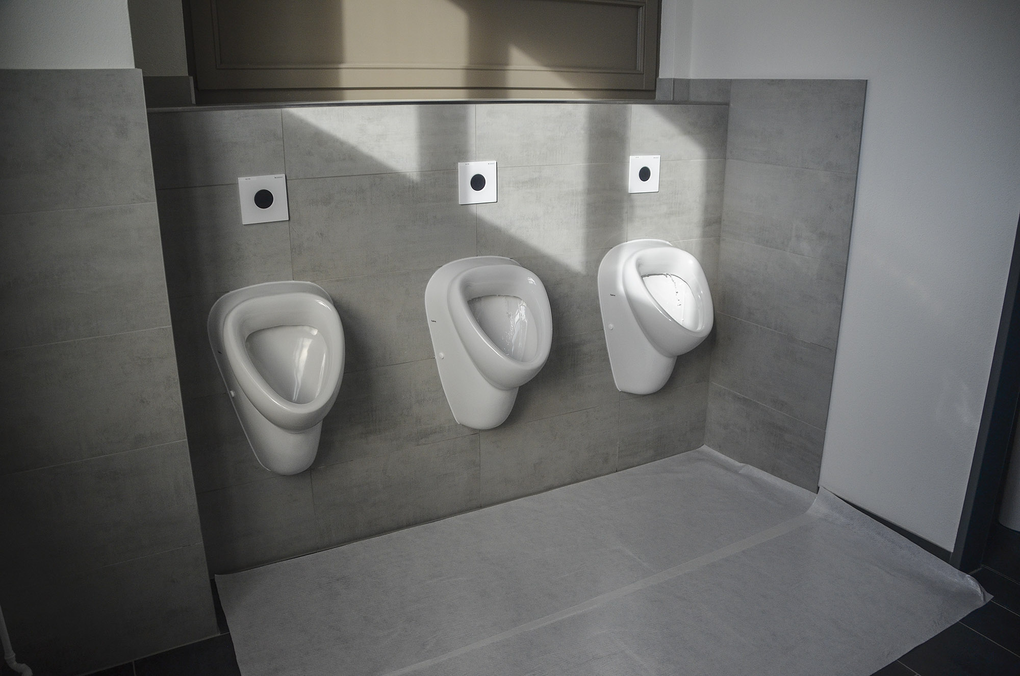 Drei nebeneinander montierte weiße Urinale an einer grauen, gefliesten Wand in einer sauberen, modernen Herrentoilette