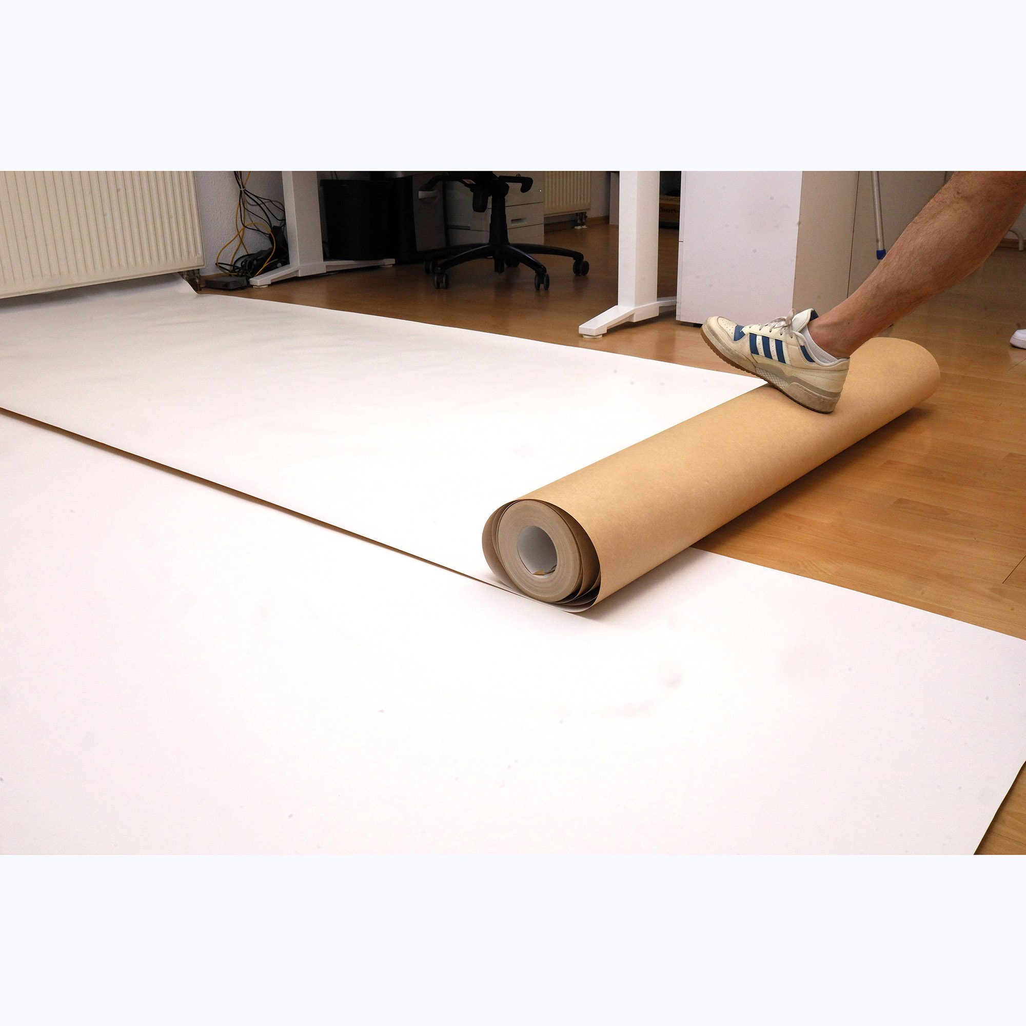 Weiße Papierrolle in brauner Hülse, die auf einem Holzboden ausgerollt wird, Fuß in Sportschuh fixiert das Ende