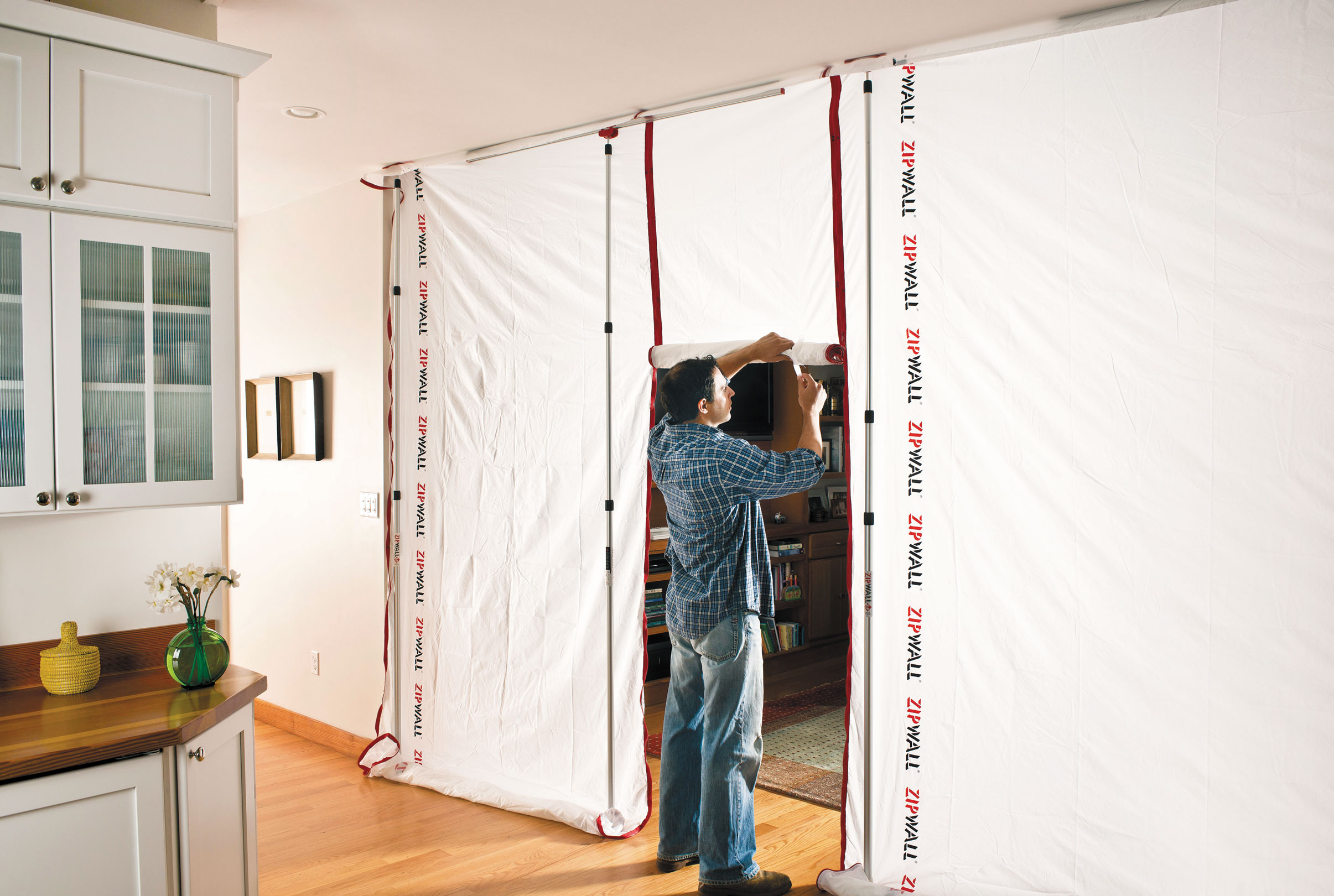Mann montiert eine ZipWall Staubschutzwand mit Reißverschluss-Tür als Trennwand im Wohnbereich, um den Raum bei Renovierungsarbeiten vor Staub zu schützen