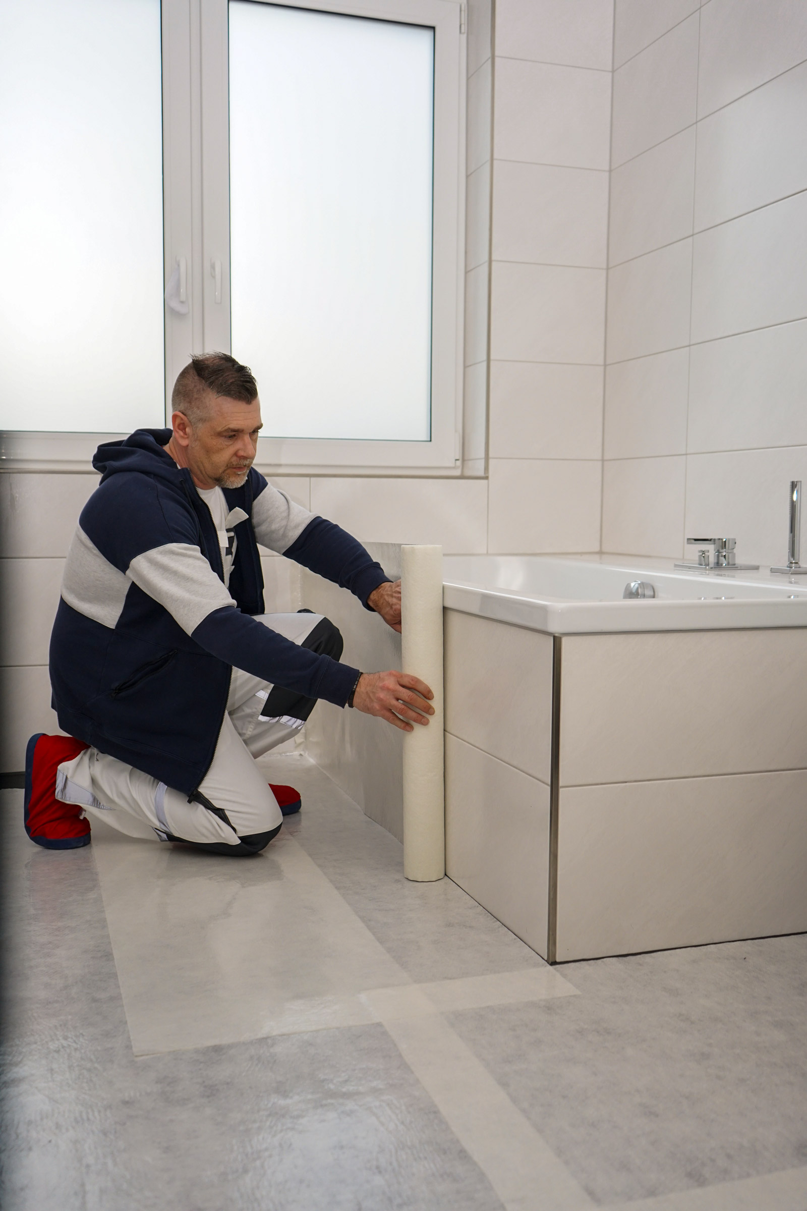 Kniender Handwerker bringt eine selbstklebende Schutzfolie im Badezimmer an der Verkleidung einer Badewanne an, um Boden und Fliesen vor Verschmutzung zu schützen.