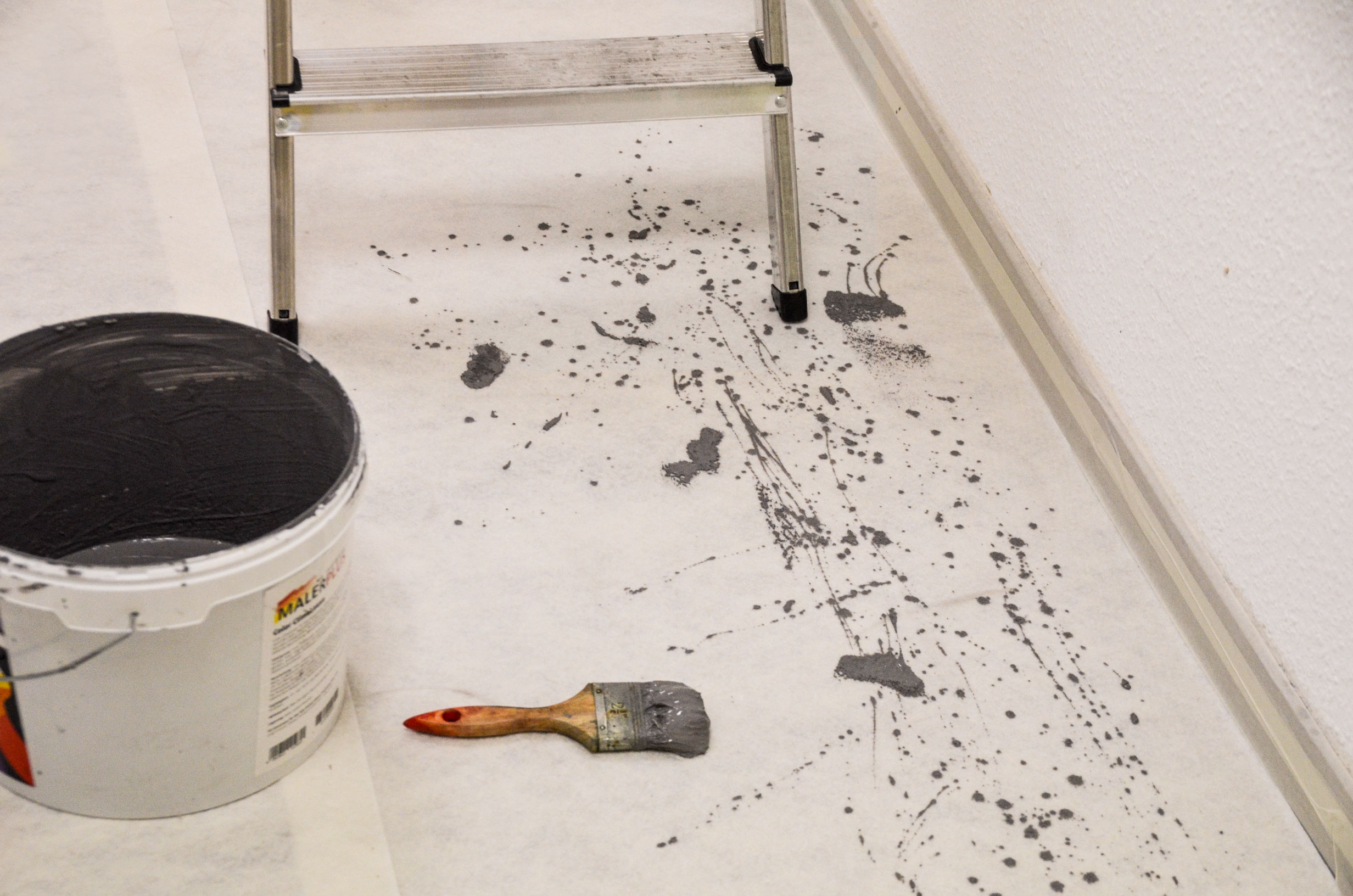 Boden mit grauen Farbspritzern vor einer Wand, daneben ein Farbeimer, ein benutzter Flachpinsel und eine Aluminiumleiter bei Renovierungsarbeiten
