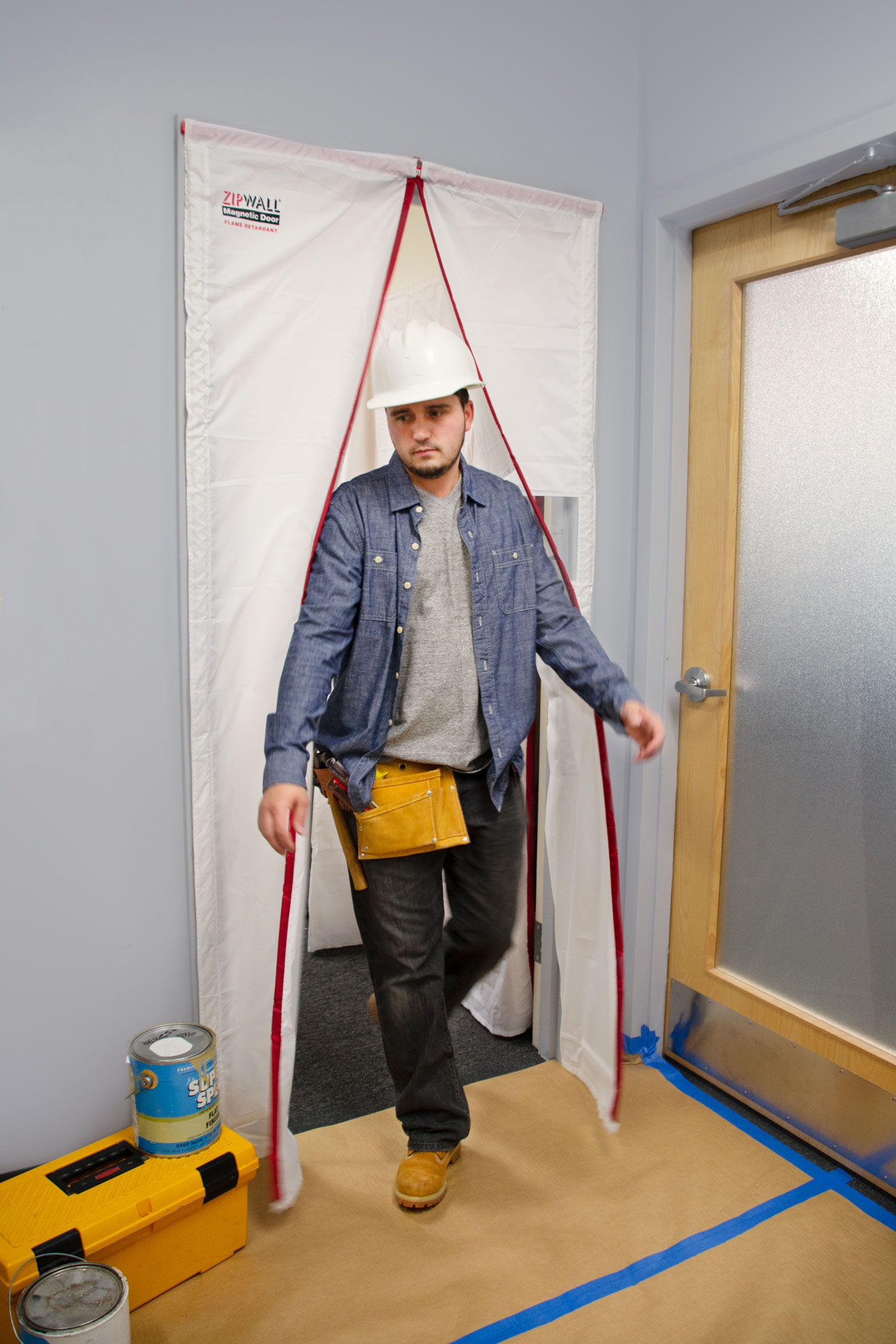 Handwerker mit Schutzhelm geht durch eine ZipWall magnetische Staubschutztür in einem innenliegenden Renovierungsbereich, Boden mit Abdeckpapier und Werkzeugkiste geschützt