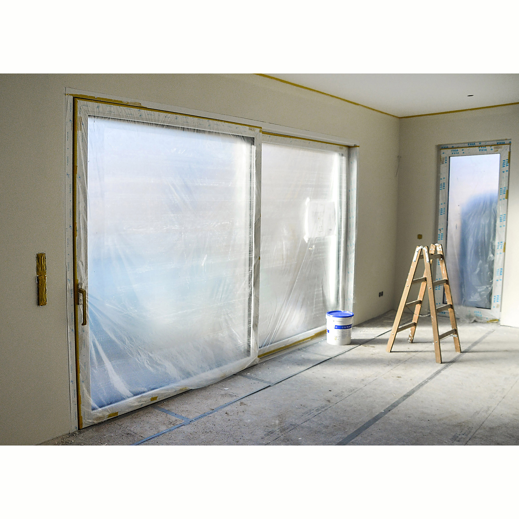 Renovierungsarbeiten in einem hellen Raum mit großen, mit Folie abgeklebteten Fenstern, einer Holzleiter und einem Farbeimer auf dem Boden