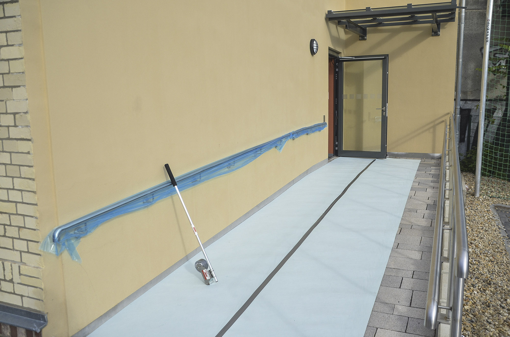 Wandabrollgerät mit langer Stange zum Abrollen von Vlies- und Folienbahnen auf einer Rampe vor einem Gebäudeeingang, geeignet zum Schutz von Böden und Wegen während Bau- oder Renovierungsarbeiten