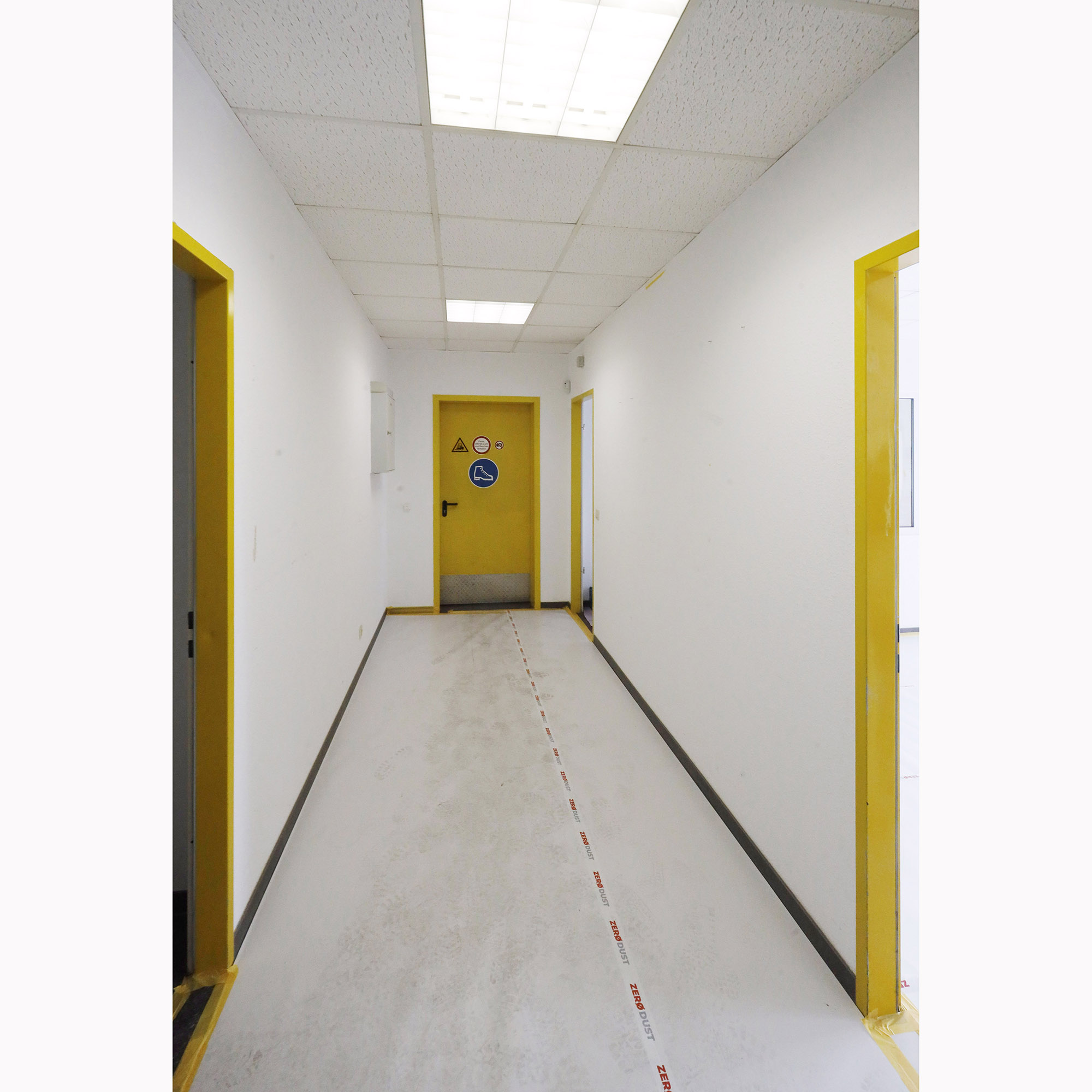 Langer, heller Flur in einem Industrie- oder Laborgebäude mit gelben Türen, weißer Decke und markiertem Sicherheitsstreifen auf dem Boden