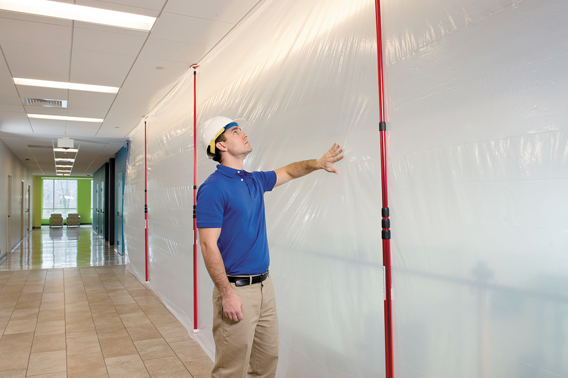 Handwerker mit Schutzhelm überprüft eine montierte Staubschutzwand aus Folie, die mit roten Teleskopstangen in einem Flur abgespannt ist