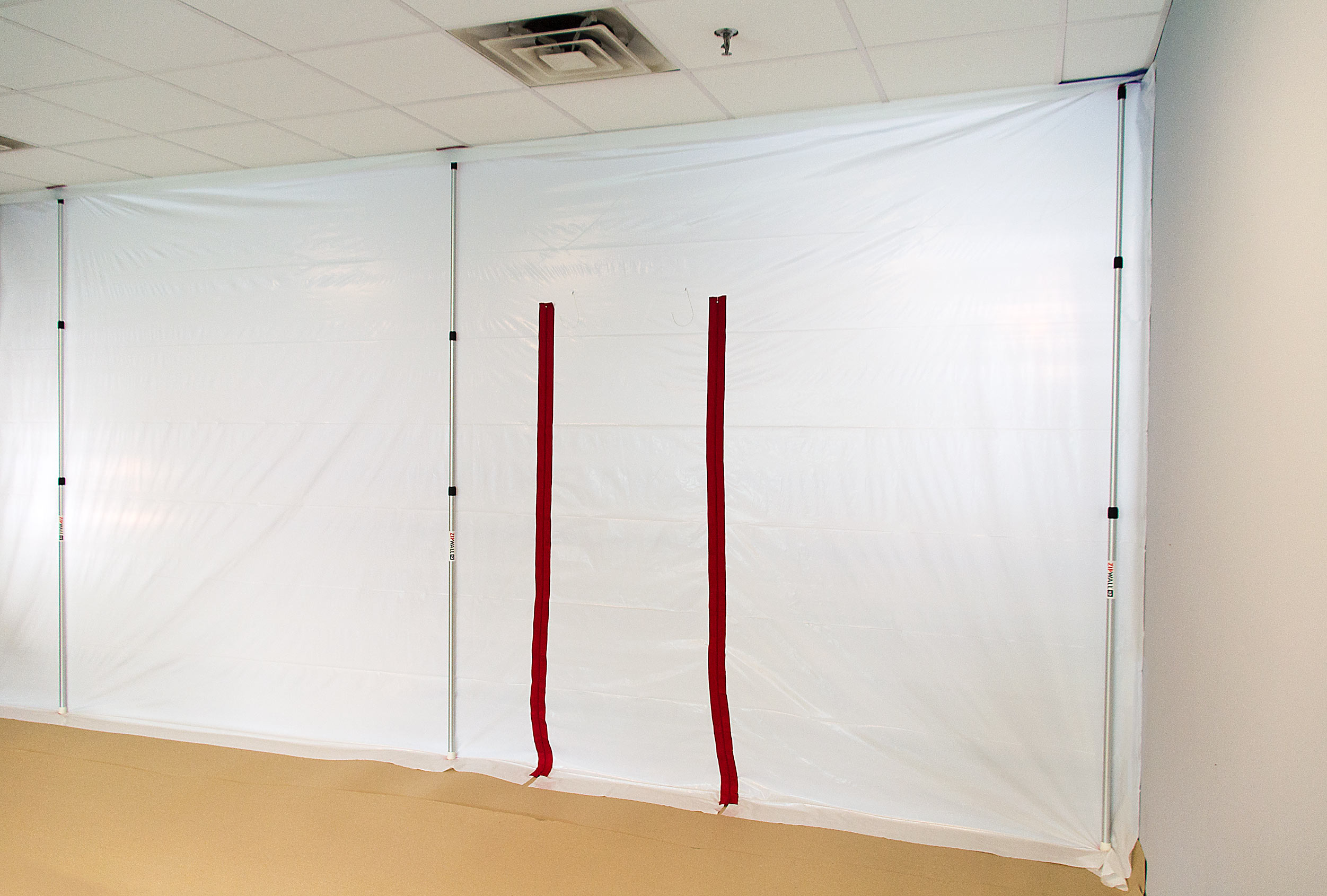Weiße Staubschutzwand aus Folie mit zwei roten Reißverschlussöffnungen und teleskopierbaren Stützstangen zur Abtrennung von Arbeitsbereichen in Innenräumen